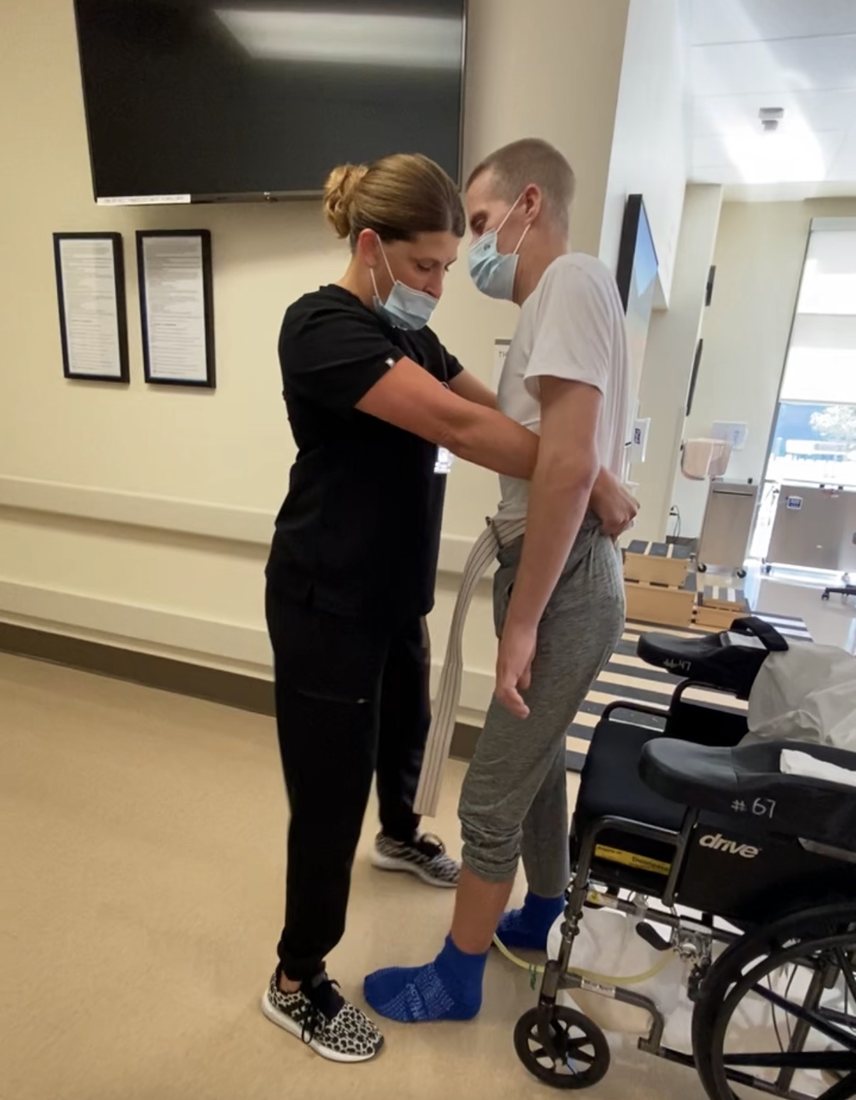 Jason standing with a physical therapist's support during inpatient rehab, an empty wheelchair beside him